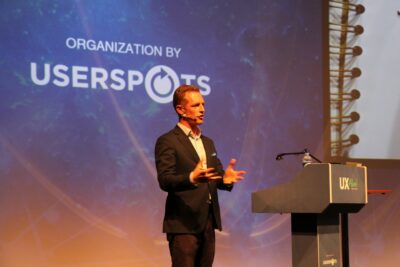 A person speaks passionately on stage beside a podium labeled UX Alive, with a backdrop displaying Organization by USERSPOTS and an image of a spiral notebook.
