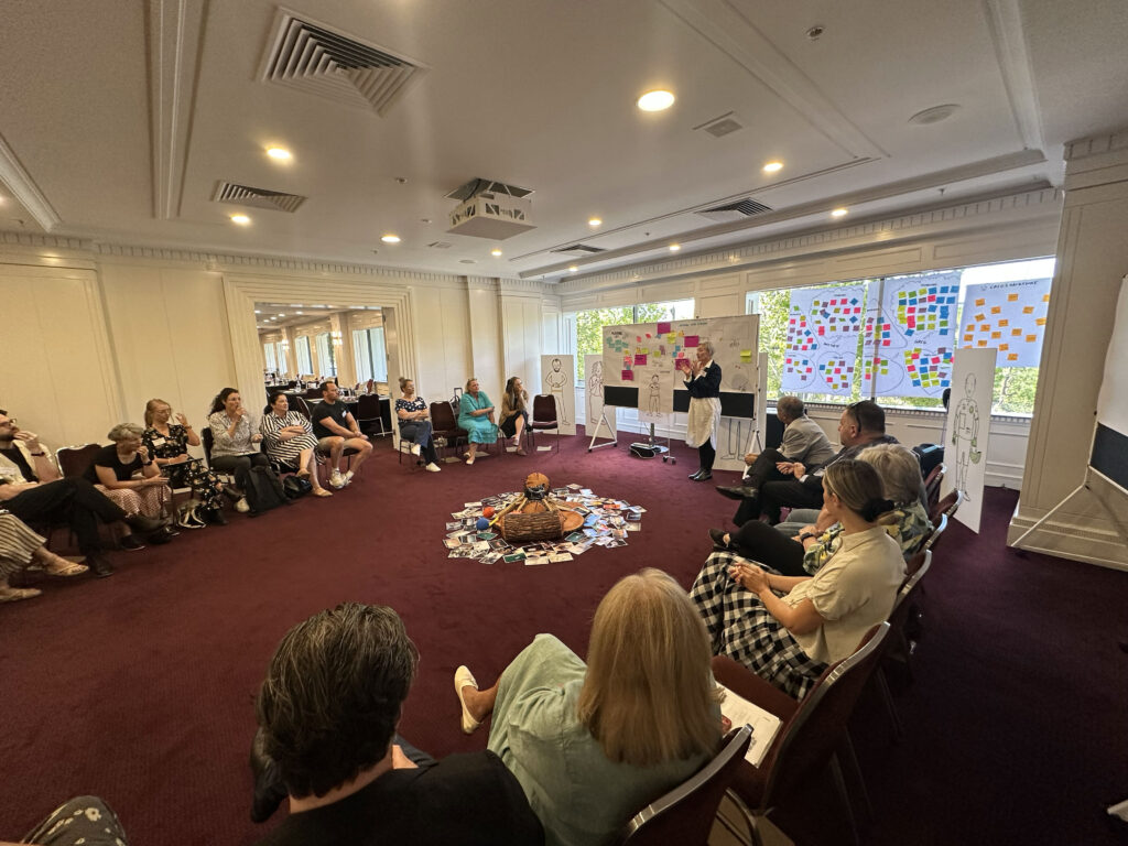 Movember Graphic Facilitation Event - group of people sitting around a pretend campfire with a facilitator leading the discussion using visual thinking tools such as a whiteboard and large foam core images of people