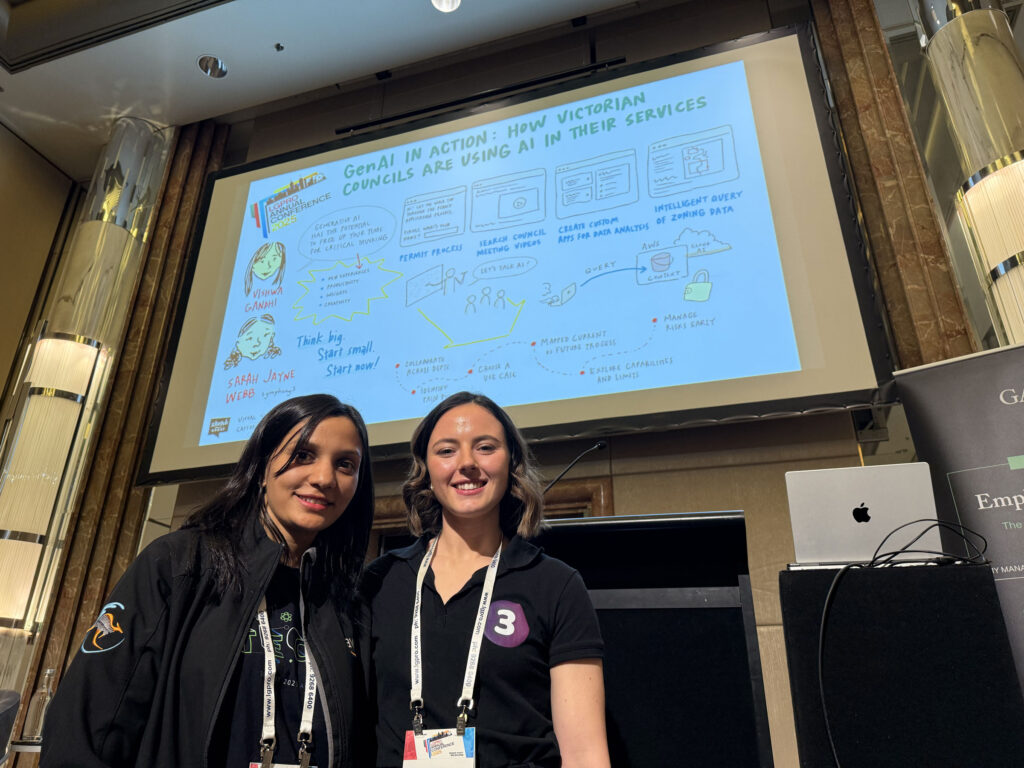 An image of 2 females from LGPro event standing in front of a graphic recording canvas broadcast on a large screen.