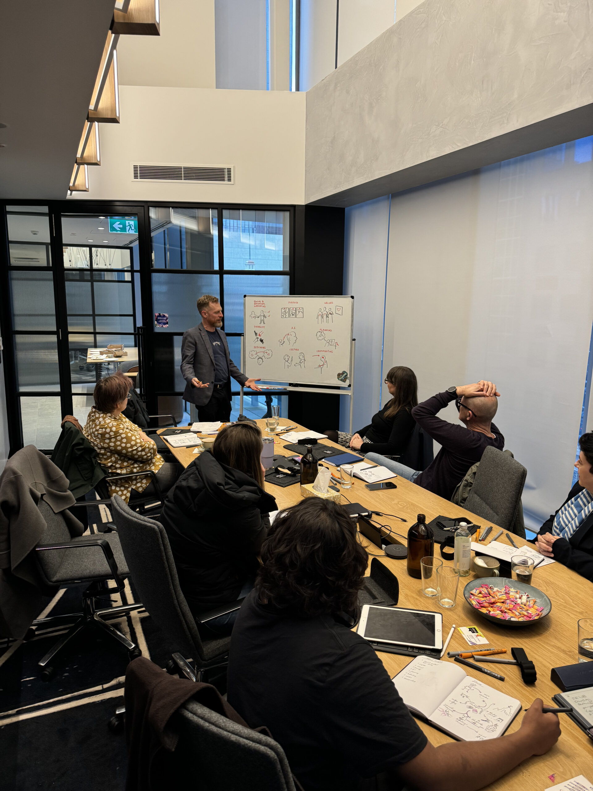 Image of people sitting around a table, watching a gentleman present on a whiteboard